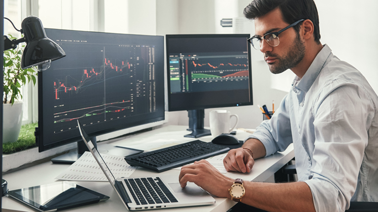 a business man sitting in front of three computer screens which have graphs displayed on the screen